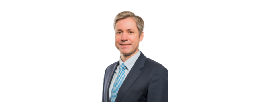 Ein mittelalter Mann ist ab dem Oberkörper zu sehen. Er guckt in die Kamera und lächelt. Er trägt ein Sakko, ein Hemd und eine Krawatte.