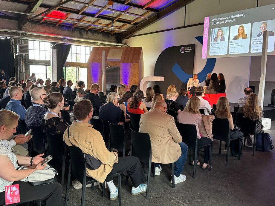 Publikum sitzt in einem Konferenzraum, auf der Bühne sitzen drei Personen und sprechen